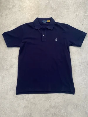 Mörkblå piké från Polo Ralph Lauren - Klassisk mörkblå pikétröja från Polo Ralph Lauren med vit broderad logga på bröstet. Modellen är custom slim fit och har korta ärmar, krage och knappar framtill. Tillverkad i mjuk bomull som känns skön mot huden.