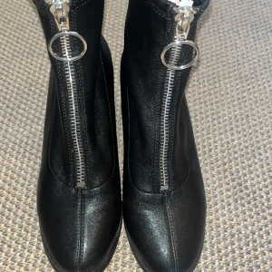 Svarta stövletter med dragkedja - Säljer ett par svarta stövletter i skinnimitation med chunky klack och rund tå. Dragkedja framtill med stor metallring som detalj. Snygga till både jeans och kjol, perfekta för höst och vinter.