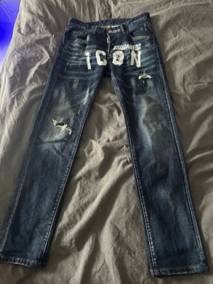 Dsquared2 ICON jeans med slitningar - Säljer ett par blå Dsquared2 ICON jeans med coola slitningar och distressed detaljer. Jeansen har vit text framtill, klassisk femficksmodell och raka ben. Materialet är robust denim i bomull och passformen är normal. Perfekta för dig som vill sticka ut med en streetwear vibe.