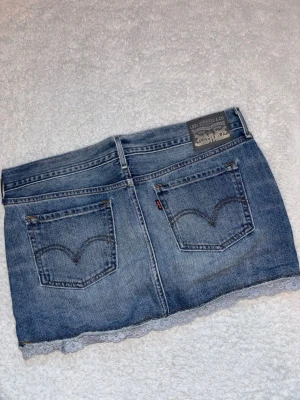 Levi's jeanskjol med spetskant - Säljer en snygg blå jeanskjol från Levi's i klassisk modell med fem fickor och bälteshällor. Kjolen har en kort passform och är dekorerad med en ljus spetskant längst ner för en extra detalj. Midjemått rakt över: 42, längd: 29 cm