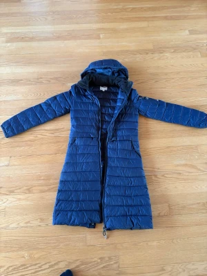 Blå lång pufferjacka från Esprit XS - Säljer en lång, blå ridjacka CRW i storlek XS. Jackan har huva, dragkedja hela vägen, fickor med dragkedja och en extra ficka på ärmen. Quiltad design och tryckknappar nertill bak för extra rörelsefrihet. Perfekt för ridning och i stallet. Inköpt på Hööks.