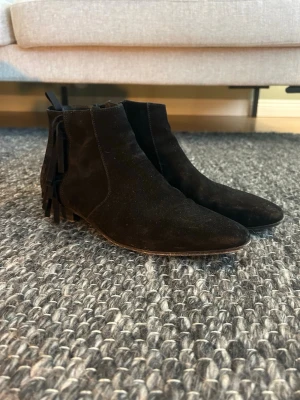 Saint laurent boots - Säljer ett par svarta boots i mocka med fransdetaljer på hälen. Skorna har rund tå, låg klack och dragkedja på insidan för enkel påtagning. Perfekta för dig som gillar stilrena men ändå unika detaljer. Innersula 27.5 cm