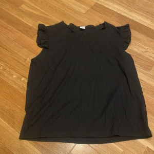 Svart ribbad topp med volangärm - Säljer en svart ribbad topp från SHEIN med volangdetaljer vid ärmöppningarna. Toppen är ärmlös och har en mjuk, stretchig känsla. 