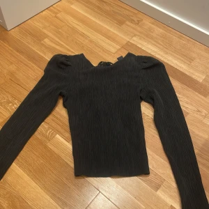 Svart långärmad topp med knyt i ryggen - Säljer en svart långärmad topp med ribbad struktur och puffiga axlar. Toppen har en snygg knytning baktill  vid urringningen och är tillverkad i ett stretchigt syntetmaterial. Passformen är normal.