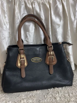 Svart handväska från Jimmy Choo - Snygg svart handväska från Jimmy Choo med bruna handtag och guldfärgade metalldetaljer. Väskan har flera fack inuti, dragkedja och spännen framtill. Tillverkad i skinn med en klassisk och stilren design som passar perfekt till din vardagslook.