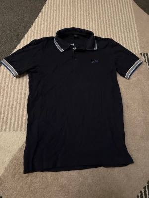 Svart pikétröja från BOSS - Klassisk svart pikétröja från BOSS med korta ärmar och krage med vita och blå ränder. Tröjan har knappar vid halsen och BOSS-logga broderad på bröstet. Perfekt för en stilren och avslappnad look. PASSAR S/M