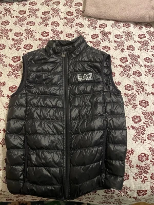 Svart dunväst från Emporio Armani EA7 - Säljer en svart dunväst från Emporio Armani EA7. Västen har quiltad design, hög krage och dragkedja framtill med EA7-logga på bröstet. Tillverkad i glansig polyester som ger en modern look. Perfekt att slänga över en hoodie eller tröja storlek s.