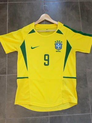 Nike Brasil Ronaldo 9 matchtröja - Gul och grön Brasilien matchtröja från Nike med nummer 9 och Ronaldo på ryggen. Tröjan har korta ärmar, gröna detaljer längs sidorna och axlarna samt det brasilianska landslagets emblem på bröstet. Perfekt för dig som älskar fotboll och vill hylla en ikon. Hör av er vid frågor och priset är inte hugget i sten 