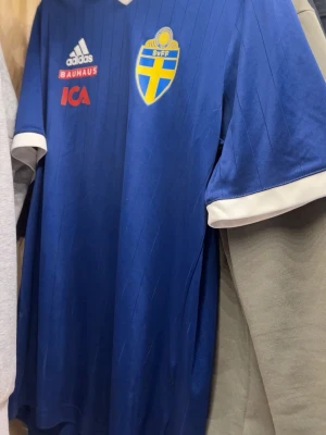 Blå Sverige fotbollströja från Adidas - Säljer en blå officiell Sverige fotbollströja från Adidas med vita detaljer och AEROREADY-tyg. Tröjan har Sveriges landslagsemblem på bröstet, Adidas-logga och sponsorer tryckta framtill. Perfekt för dig som vill visa ditt stöd på matchen!