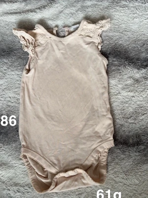 Aprikosfärgad ärmlös body med spetsdetaljer - Supersöt aprikosfärgad body från Newbie med ärmlös design och fina spetsvolanger på axlarna. perfekt för varma dagar. Knäppning bak i nacken för enkel påklädning. Enkel och stilren look med romantiska detaljer.🌸Fint använt skick. Storlek 86