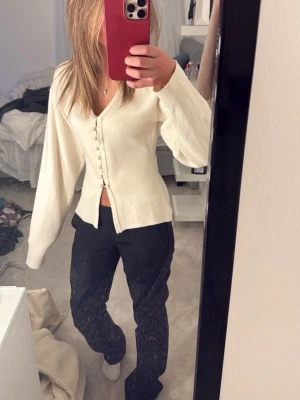 Vit kofta med knappar och v-ringning - Säljer en stilren vit kofta med v-ringning och dekorativa knappar framtill. Koftan har långärmad design och lätt croppad passform som ger en snygg siluett. 