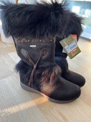 Mörkbruna pälsboots från Tecnica - Säljer ett par mörkbruna Italienska kalvläder boots från Tecnica med äkta  päls och dekorativa detaljer upptill. Skorna har rund tå, platt sula och snörning på sidan. Utsidan är täckt av päls och mocka, vilket ger en riktigt mysig vinterlook. Perfekta för kalla dagar! Stövlarna är helt nya, kapparna sitter kvar. Nypris 3.600kr. 