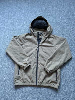 Beige/grön vindjacka från Wayfarer Young - Snygg beige vindjacka med huva från Wayfarer Young. Jackan har hel dragkedja framtill, två sidofickor med dragkedja och elastiska muddar vid ärmslut och nederkant. Perfekt för blåsiga dagar och enkel att matcha med olika outfits.