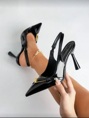 Svarta lackpumps med gulddetalj - Snygga svarta pumps i lack med spetsig tå och hög klack. Skorna har en öppen häl och en elegant guldfärgad spänndetalj framtill. Perfekt för dig som gillar att sticka ut med stilrena och trendiga skor. Materialet är glansigt syntet och designen är riktigt chic. Använda endast en utgång 