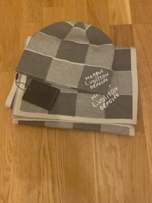 Gråvit rutig Louis Vuitton set - Snygg väst från Louis Vuitton i grått och vitt med rutigt mönster. Västen har broderad text 'Marque L. Vuitton Déposé' framtill och är tillverkad i ett mjukt stickat material. Perfekt för dig som vill ha en exklusiv och trendig look.