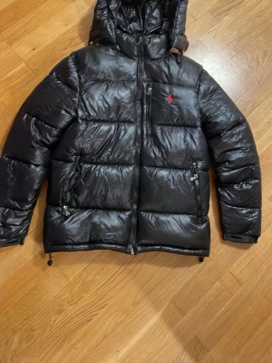 Svart pufferjacka från Polo Ralph Lauren - Säljer en svart pufferjacka från Polo Ralph Lauren med glansig finish och röd logga på bröstet. Jackan har huva, dragkedja framtill och två snedställda fickor med dragkedja. Perfekt för kalla dagar och riktigt snygg streetstil.