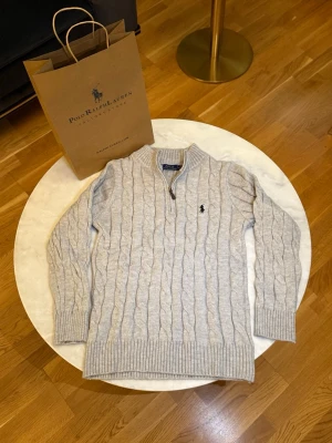 Beige stickad half zip tröja Polo Ralph Lauren - Säljer en beige stickad tröja från Polo Ralph Lauren i storlek M. Tröjan har ett klassiskt kabelstickat mönster, ribbad krage och muddar samt en half zip-dragkedja framtill. Ikoniska Polo-loggan broderad på bröstet. Perfekt för kyliga dagar.