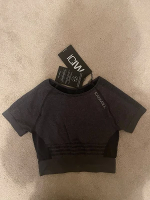 ICIW träningströja  - Bilderna visar en mörkgrå kortärmad topp från ICANIWILL. Produkten har ribbade detaljer och är tillverkad i ett stretchigt syntetmaterial, perfekt för träning.