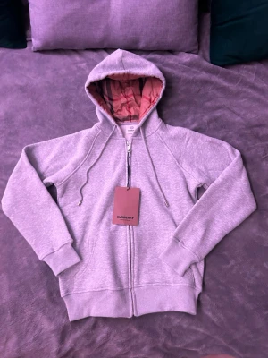 Burberry’s Zip-Hoodie - Visst, här är en beskrivning du kan använda för din Vinted-artikel:  Burberry Zip-Hoodie med Rutigt Foder  Säljer en helt ny Burberry-zip-hoodie, oanvänd med etiketter kvar. Denna hoodie är tillverkad i mjukt, grått bomullstyg och har ett ikonisk rutigt foder i huvan. Perfekt för att lägga till en touch av lyx till din vardagsgarderob.  *   Skick: Helt ny med etiketter *   Färg: Grå *   Detaljer: Burberry-rutigt foder i huvan, dragkedja framtill, fickor *   Storkel: S