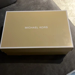 Svarta slip-on sneakers från Michael Kors köpte skorna i Dubai  - Snygga slip-on sneakers från Michael Kors i svart med diskret logomönster. Skorna har vit platt sula med Michael Kors-tryck på sidan och under sulan. Ovandelen är i syntetmaterial med en stilren look, perfekt till jeans eller kjol.
