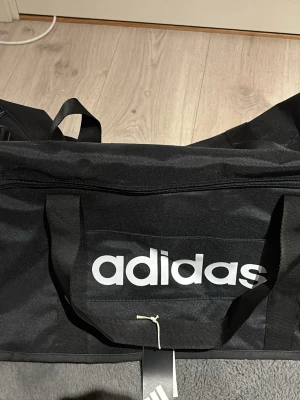 Svart Adidas duffelbag stor - Säljer en stor svart duffelbag från Adidas med tydlig vit logga på sidan. Väskan har justerbar axelrem, bärhandtag och dragkedja upptill. Perfekt för gymmet eller resan, rymmer mycket och är riktigt stilren. Väskan är helt ny och är aldrig använd, säljer då jag ej fått nån användning för den, nypris är 499, mitt pris 299, mvh
