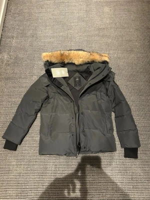 Mörkgrå pufferjacka med fuskpäls - Säljer en mörkgrå pufferjacka från Moose Knuckles med avtagbar huva och fuskpälsdetalj. Jackan har dragkedja och knappar framtill, ribbade muddar vid ärmslut och flera praktiska fickor. Perfekt för kalla vinterdagar.