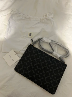 Svart axelväska från By Malene Birger - Snygg svart axel fäska från By malene Birger med graf tap pin trutig mönster. Väskan har guldfärgade detaljer, dragkedja och avtagbar axelrem. Kommer med dustbag och tag kvar. Perfekt storlek för det viktigaste och stilren look.