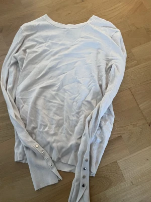 Vit långärmad tröja med knappar från Zara  - Säljer en stilren vit långärmad tröja med snygga knappdetaljer vid ärmsluten. Tröjan har en enkel, klassisk passform och är gjord i mjukt material som känns skönt mot huden. Perfekt att matcha med jeans eller kjol för en clean look.