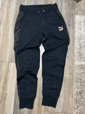 Svarta mjukisbyxor från Puma - Svarta mjukisbyxor från Puma med guldfärgad logga på vänster sida. Byxorna har ribbade muddar vid benslut, snörning i midjan och diskreta ränder i materialet längs sidorna. Perfekta för chill eller träning.