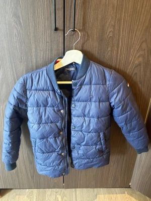 Blå quiltad bomberjacka Tommy Hilfiger - Snygg blå quiltad bomberjacka från Tommy Hilfiger med ribbade muddar och krage. Jackan har tryckknappar framtill och diskret logga på bröstet samt ärmen. Perfekt för kyliga dagar och ger en cool look.