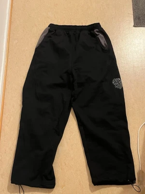 Svarta mjukisbyxor från Merta - Svarta mjukisbyxor från Murtra med grå detaljer vid fickorna och en vit grafisk logga på benet och bakfickan. Byxorna har elastisk midja och snörning vid bensluten för extra stilpoäng. Oanvänd, perfekt skick.