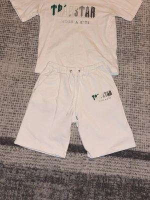 Vita Trapstar shorts med snörning och palm angels tröja - Säljer ett par vita shorts från Trapstar i storlek M. De har elastisk midja med snörning och logga tryckt på vänster ben. Materialet är mjukt och känns som bomull, perfekt för chill dagar. Klassisk loose fit och fickor på sidorna.