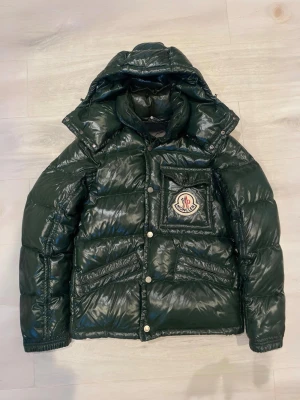 Mörkgrön Moncler pufferjacka - Säljer en mörkgrön pufferjacka från Moncler med glansig finish och stor logga på bröstet. Jackan har huva, dragkedja och knappar framtill samt flera fickor. Perfekt för kalla dagar och riktigt snygg till streetwear-stilen.