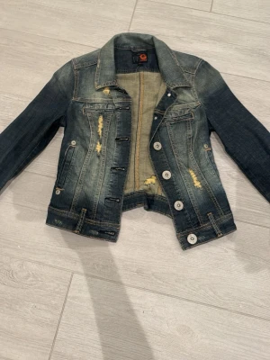 Blå jeansjacka från Guess - Säljer en klassisk blå jeansjacka från Guess med slitna detaljer och snygga kontrastsömmar. Jackan har knappar framtill, två bröstfickor och långa ärmar. Perfekt för dig som gillar en avslappnad och trendig look.