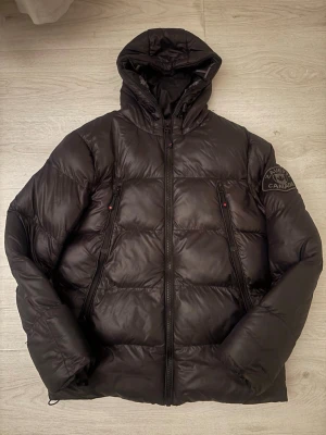 Svart pufferjacka från Zavetti Canada - Svart pufferjacka från Zavetti Canada med huva och feta logodetaljer på insidan. Jackan har två dragkedjeförsedda fickor framtill och quiltad design. Perfekt för kalla dagar och har en riktigt schysst streetstil.