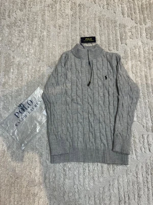 Grå kabelstickad tröja Polo Ralph Lauren - Kabelstickad grå tröja från Polo Ralph Lauren med halv dragkedja och hög krage. Klassisk broderad logga på bröstet. Tröjan är långärmad och har ribbade muddar och nederkant. Perfekt för kyliga dagar och snygg till jeans.