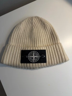Vit ribbstickad mössa från Stone Island - Säljer en snygg vit ribbstickad mössa från Stone Island med den klassiska logotypen framtill. Mössan är tillverkad i mjuk ull och har en uppvikt kant. Perfekt accessoar för kyliga dagar och ger en clean look.