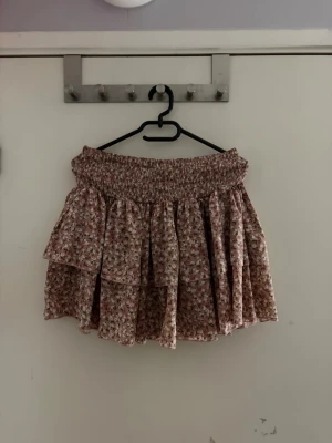 Blommig volangkjol i rosa toner - Supersöt kort volangkjol med blommigt mönster i rosa och vita nyanser. Kjolen har bred smock i midjan för skön passform och dubbla lager volanger som ger en luftig och lekfull look. Perfekt för sommarens alla dagar!