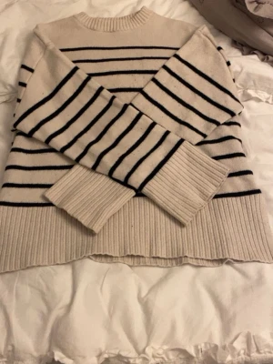 Randig beige stickad tröja - Mysig beige stickad tröja med svarta horisontella ränder. Tröjan har långa ärmar och breda ribbade muddar vid ärmslut och nederkant.