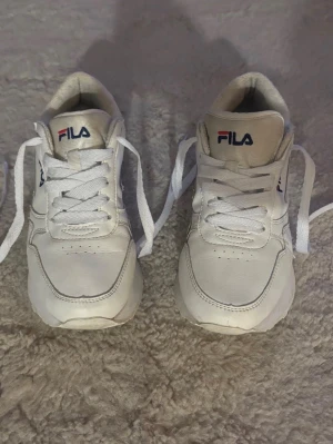 Vita Fila sneakers - Säljer ett par klassiska vita Fila sneakers med platt sula och rund tå. Skorna har snörning och Fila-logga på både plös och häl. Tillverkade i syntetmaterial med diskreta sömmar och enkel design som passar till det mesta.