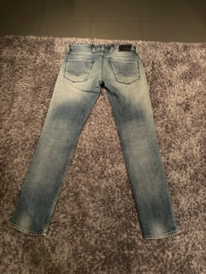 Replay Anbass ljusblå jeans, stl 29 - Säljer ett par Replay Anbass jeans i ljusblå tvätt med snygga slitningar och klassiska fem fickor. Jeansen har normal passform och raka ben, med Replay-logga på bakfickan och läderpatch i midjan. Perfekt för dig som gillar stilrena och bekväma jeans. Jätte fina verkligen! Storlek 29/32