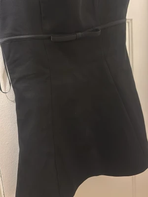 Svart kort klänning PrettyLittleThing - Snygg svart kort klänning från PrettyLittleThing med breda axelband och en söt liten rosett i midjan. Klänningen har figurnära passform och dragkedja i sidan för enkel påklädning. 