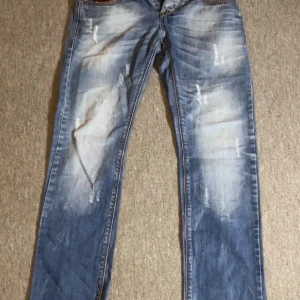 Blå slitna jeans med raka ben - Säljer ett par blå jeans med slitningar och ljusa tvättade partier. Jeansen har raka ben och klassisk femficksmodell. Materialet är jeans och de har en cool, avslappnad look som passar perfekt till sneakers eller boots.