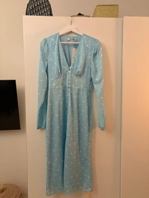 Ljusblå långklänning från Adoore - Supersöt heltny långklänning från Adoore i ljusblått med små vita blommor. Klänningen har v-ringning, långa ärmar och knappar framtill. Prislapp fina kvar, och har aldrig använt den. Modellen heter Paris Dress Pool Blue. 