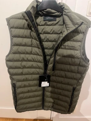 Grön dunväst från Peak Performance - Säljer en snygg, quiltad dunväst från Peak Performance i färgen Pine Needle. Västen har hög krage, svarta dragkedjor och två sidofickor. Perfekt att slänga över en hoodie eller tröja för extra värme. Klassisk logga på bröstet och stilren look. Knappt använd då den inte passar längre 