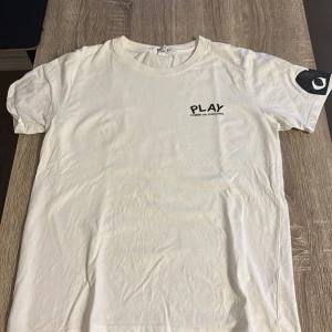 Vit Comme des Garçons Play t-shirt - Säljer en vit t-shirt från Comme des Garçons Play i storlek S. T-shirten har korta ärmar, rund hals och ett svart tryck med PLAY-loggan på bröstet. På ena ärmen finns det ikoniska svarta hjärtat med ögon. Materialet är mjuk bomull.