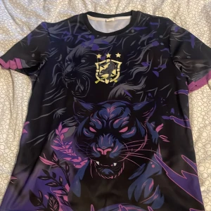 Svart och lila t-shirt med pantermotiv - Unik t-shirt med ett stort pantermotiv i lila och rosa toner på svart bakgrund. Framsidan har ett tryck med panter och ett emblem med stjärnor, medan baksidan har coola klösmärken. T-shirten är kortärmad och gjord i ett glansigt polyestermaterial som känns sportigt.