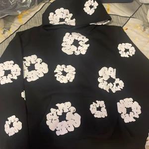 Svart hoodie från Denim Tears med blommor - Svart hoodie från Denim Tears med stora vita blommönster över hela tröjan. Klassisk huva och känguruficka framtill. Snygg och unik streetwear-stil som sticker ut i mängden.