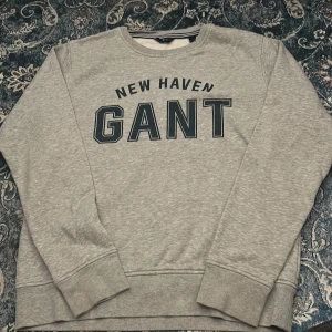 Grå sweatshirt från GANT med tryck - Säljer en grå sweatshirt från GANT med stort svart tryck 'NEW HAVEN GANT' på bröstet. Tröjan har rund halsringning med randig detalj på insidan och är tillverkad i mjuk bomullsmix. Perfekt för en chill och stilren look.