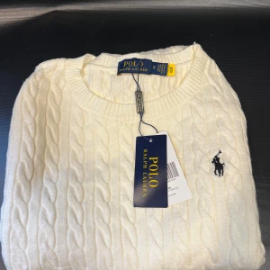 Vit kabelstickad tröja - Vit kabelstickad tröja med klassisk rund halsringning och broderad logga på bröstet. Tröjan har en stilren look och är tillverkad i mjukt material som passar perfekt för lager-på-lager under kyliga dagar.